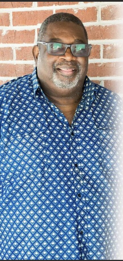 Bishop Dr. William BGK Harris smiling in a blue patterned shirt in front of a brick wall